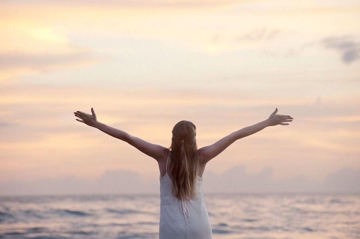 Frau in Harmonie mit der Natur Eine Frau mit ausgestreckten Händen, die vor einem weiten Ozean steht und sich mit der Natur verbindet. Diese Pose symbolisiert Freiheit, Entspannung und das Streben nach ganzheitlichem Wohlbefinden.