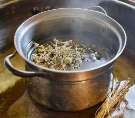Kräuter im heißen Wasserbad für Yoni Steaming Kräuter werden in ein heißes Wasserbad gegeben, um den Dampf mit ihren wohltuenden Wirkstoffen anzureichern, ideal für Yoni Steaming-Anwendungen.